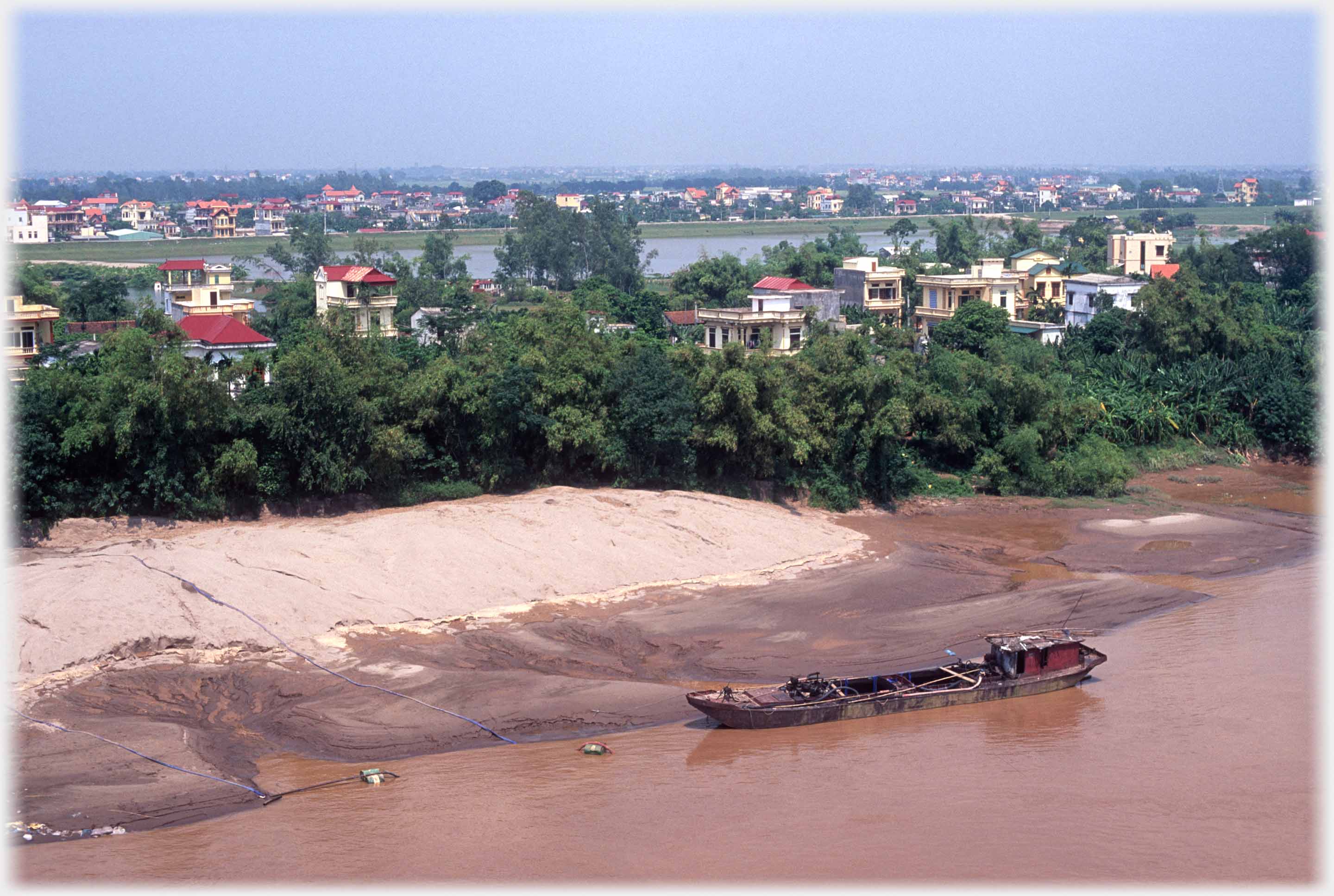 Barge at river bank which is tree covered with houses among the the trees.