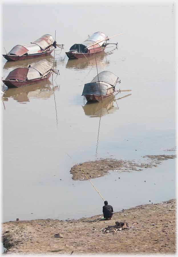 Man sitting fishing at side of river with houseboats beyond.
