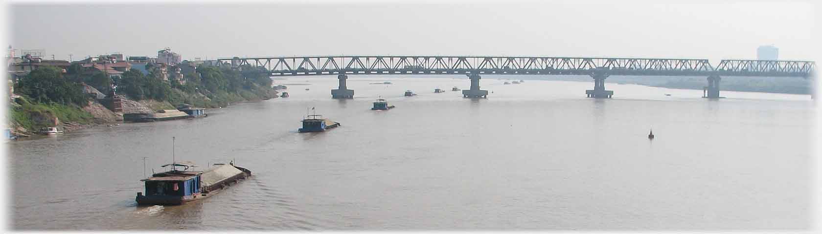 Bridge on piers over river with line of barges moving under it.