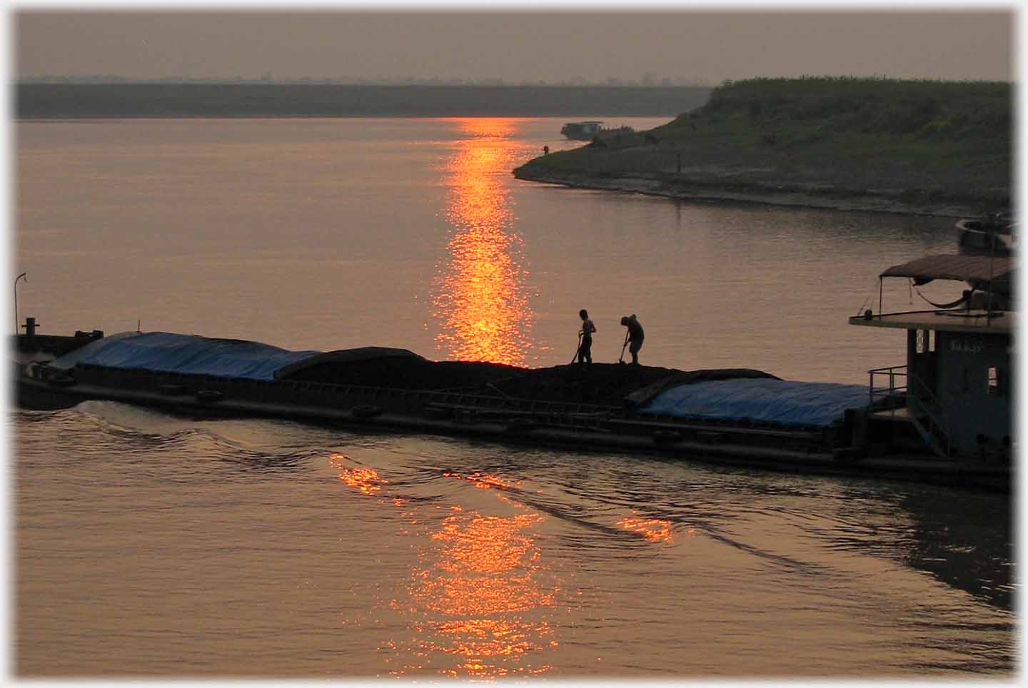 Two men working on heaps of material on barge, making small waves, with sun setting beyond.