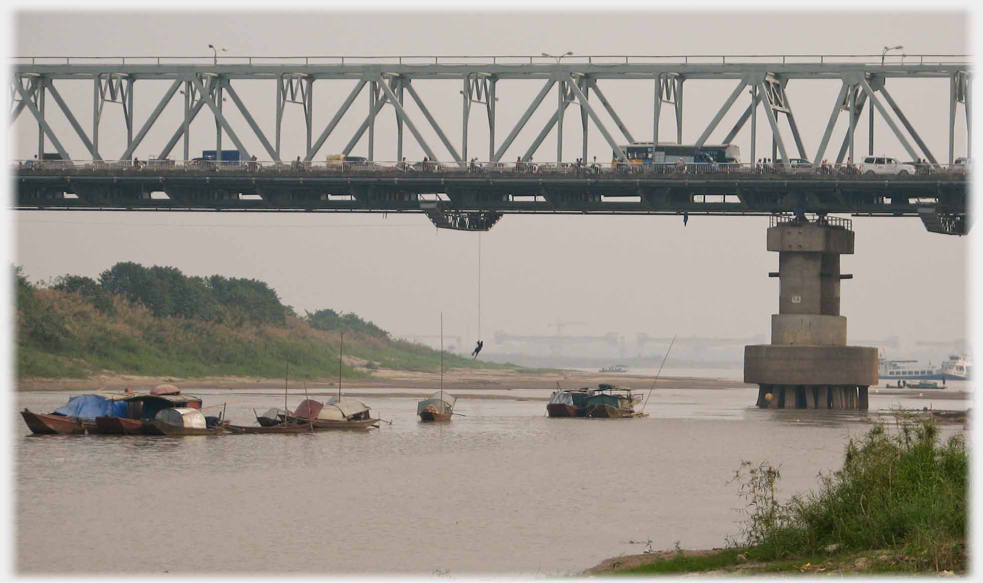 Bridge busy with traffic from which a rope is hanging with person on end of it, houseboats and river below.