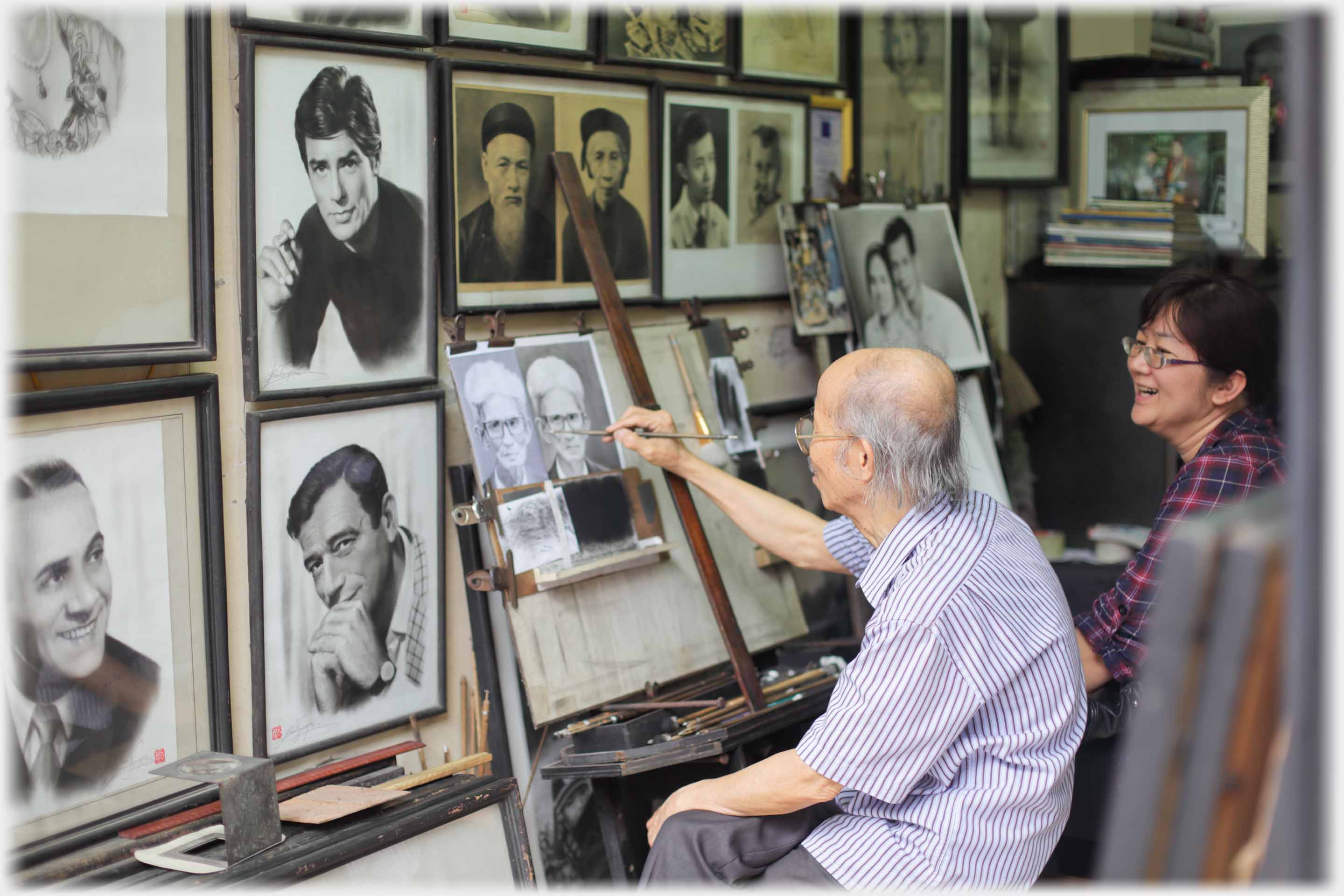 Older man painting small portrait from photograph, wall covered in photographs, woman watching.
