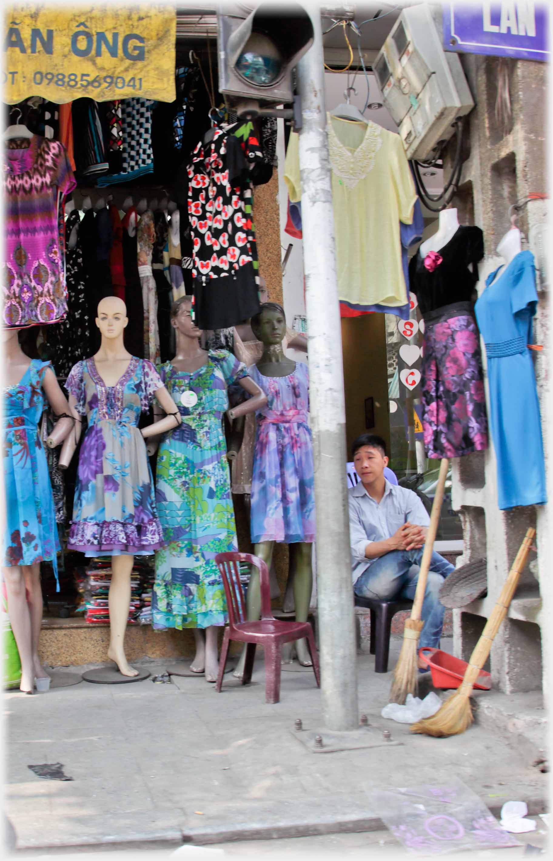 Manikins in light dresses, one with leg missing standing behind man sitting in chair.
