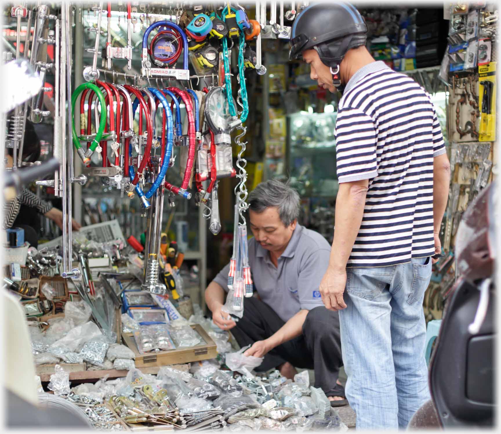 Vendor squatting filling small bag from boxes, crash-helmeted customer watching.