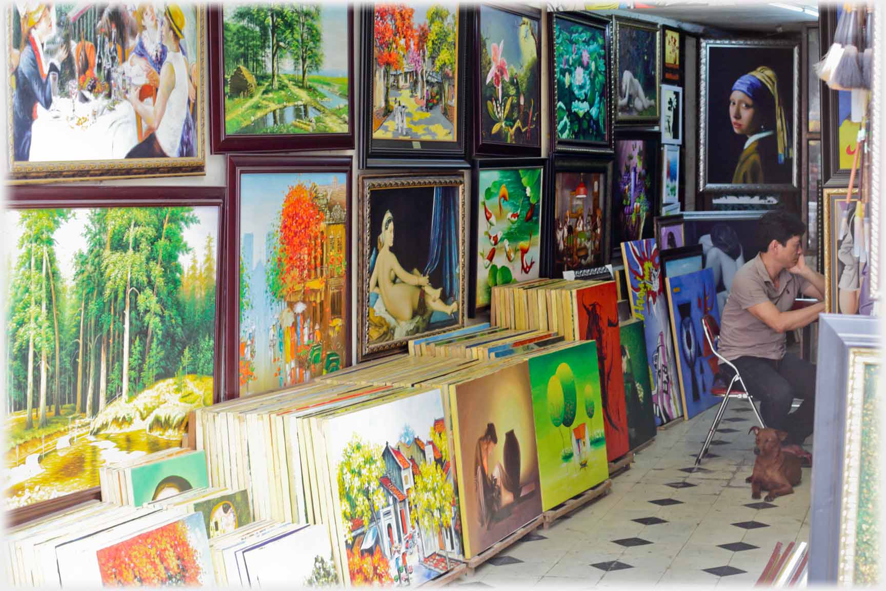 Looking into wall of shop covered in paintings, paintings stacked along floor and man sitting.