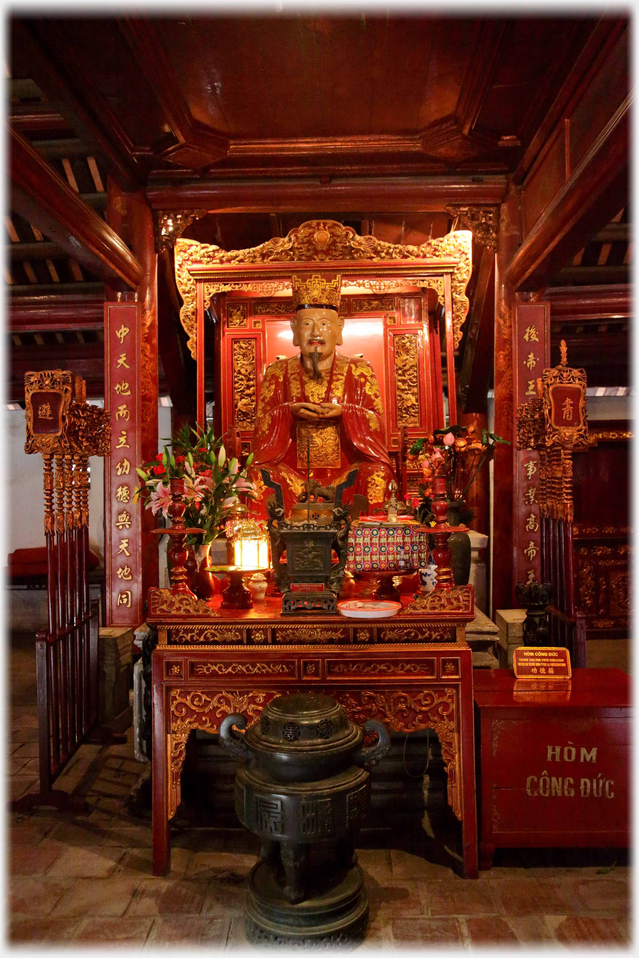 Seated red and gold figure surrounded by plants and symbolic objects.