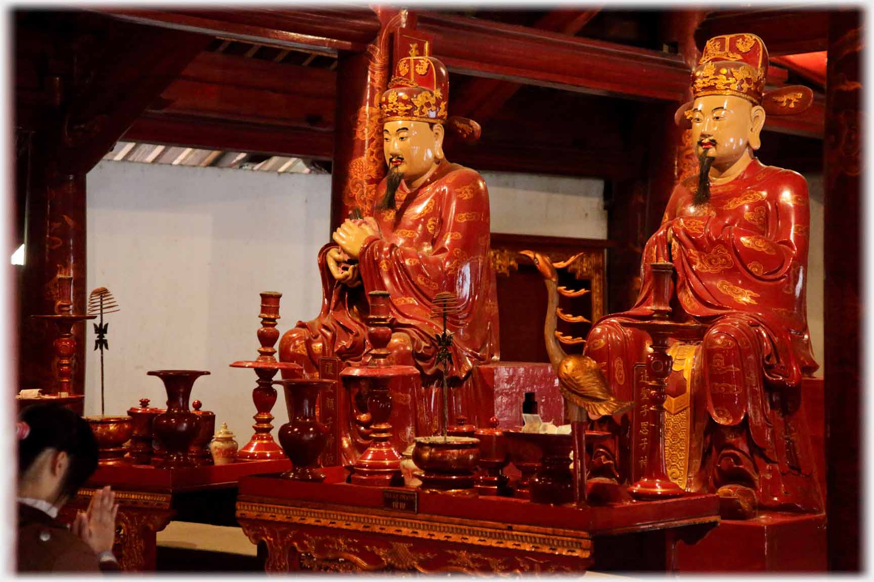 Two red and gold seated figures looking left, praying hands visible.