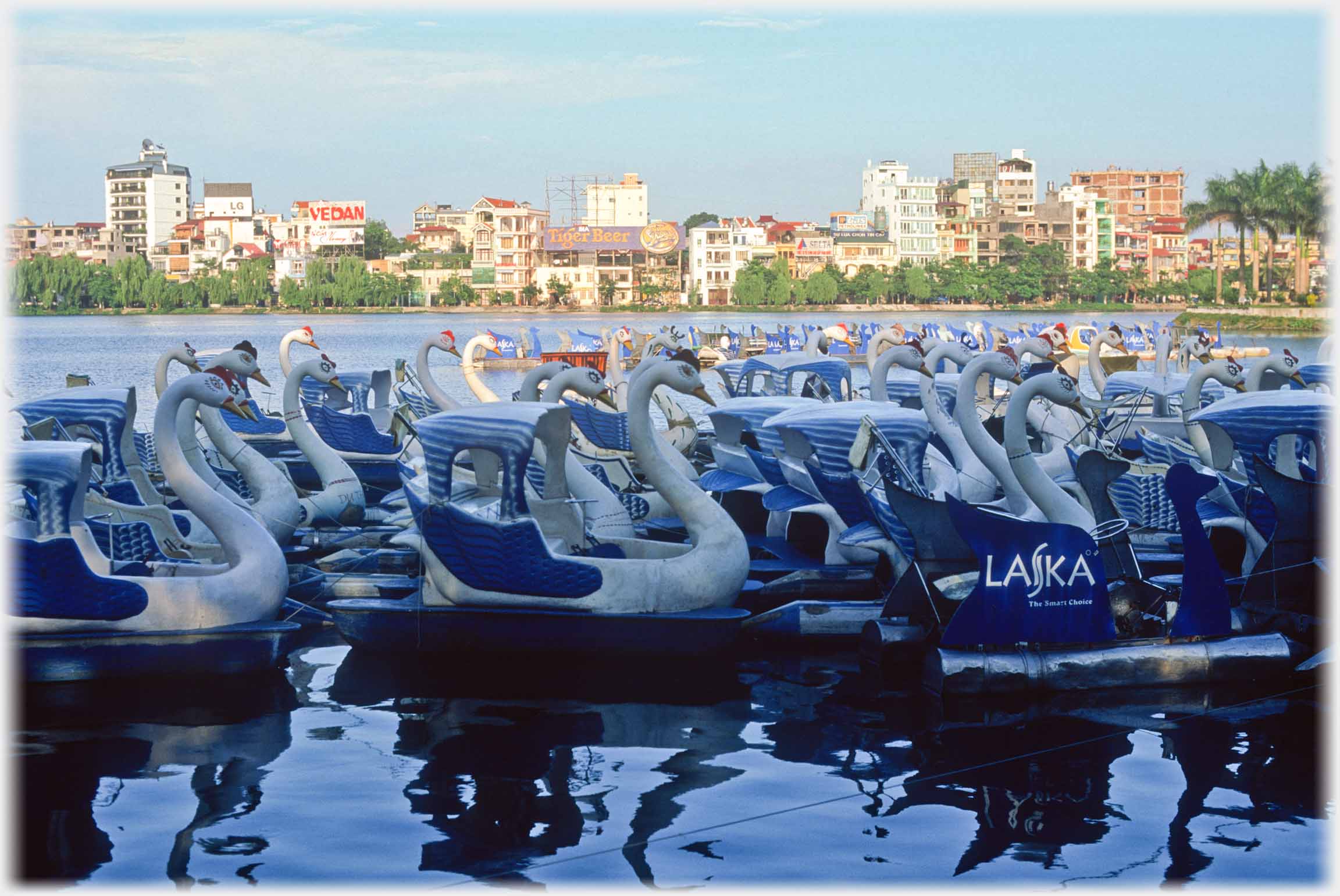 Packed area of pedalos in shadow, bright sun on houses across lake.