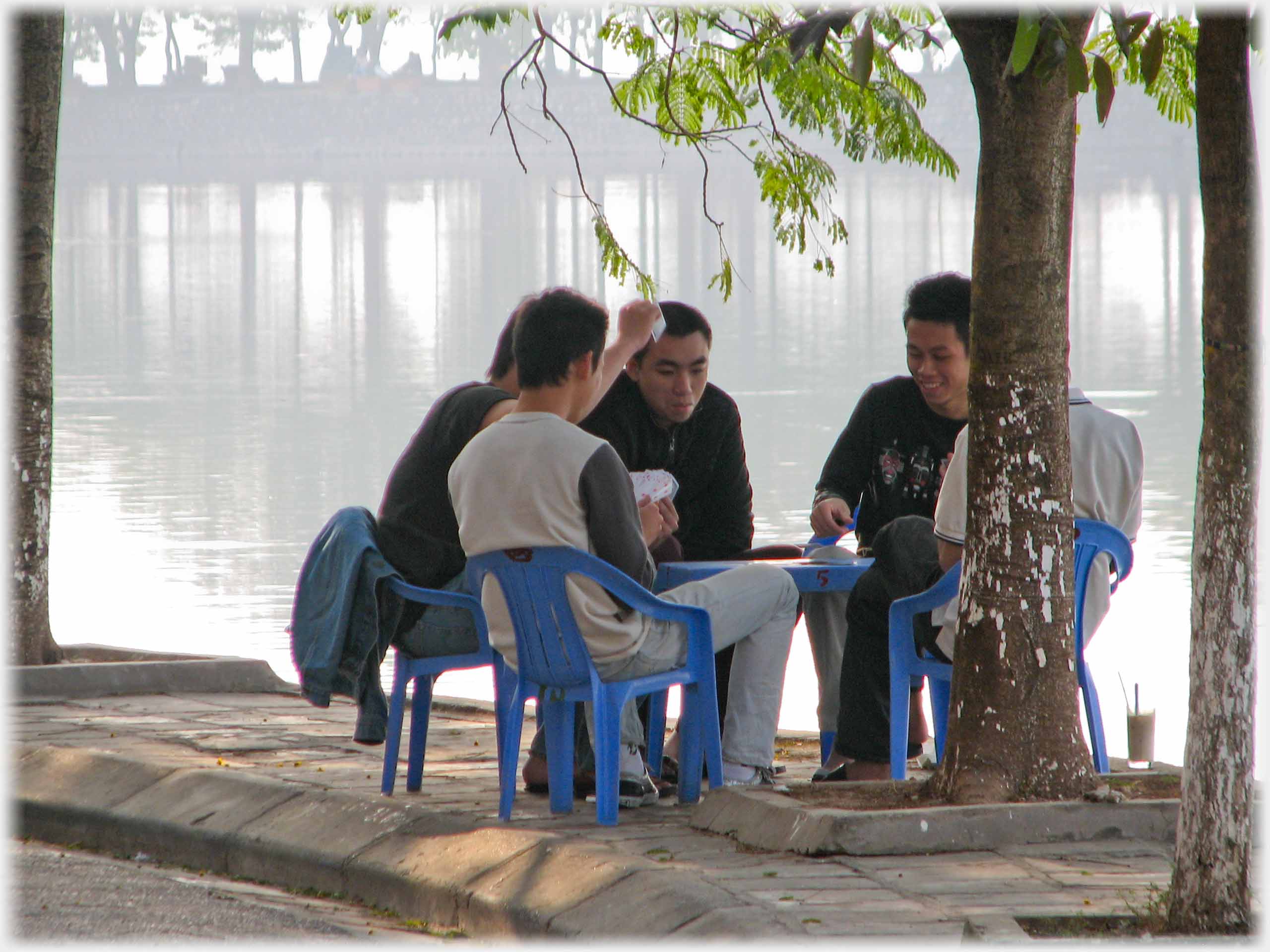 Five men on chairs round table playiing cards by lake.