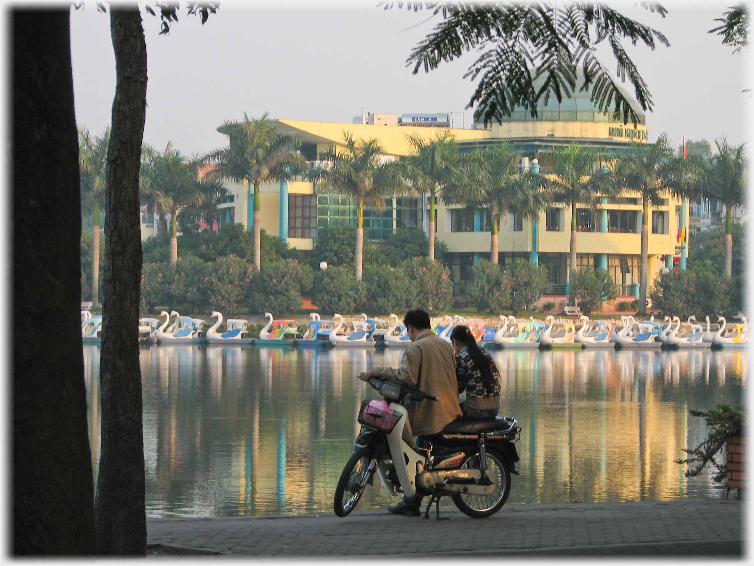 Couple sitting sidesaddle on motorbike looking across at pedalos and restaurant.