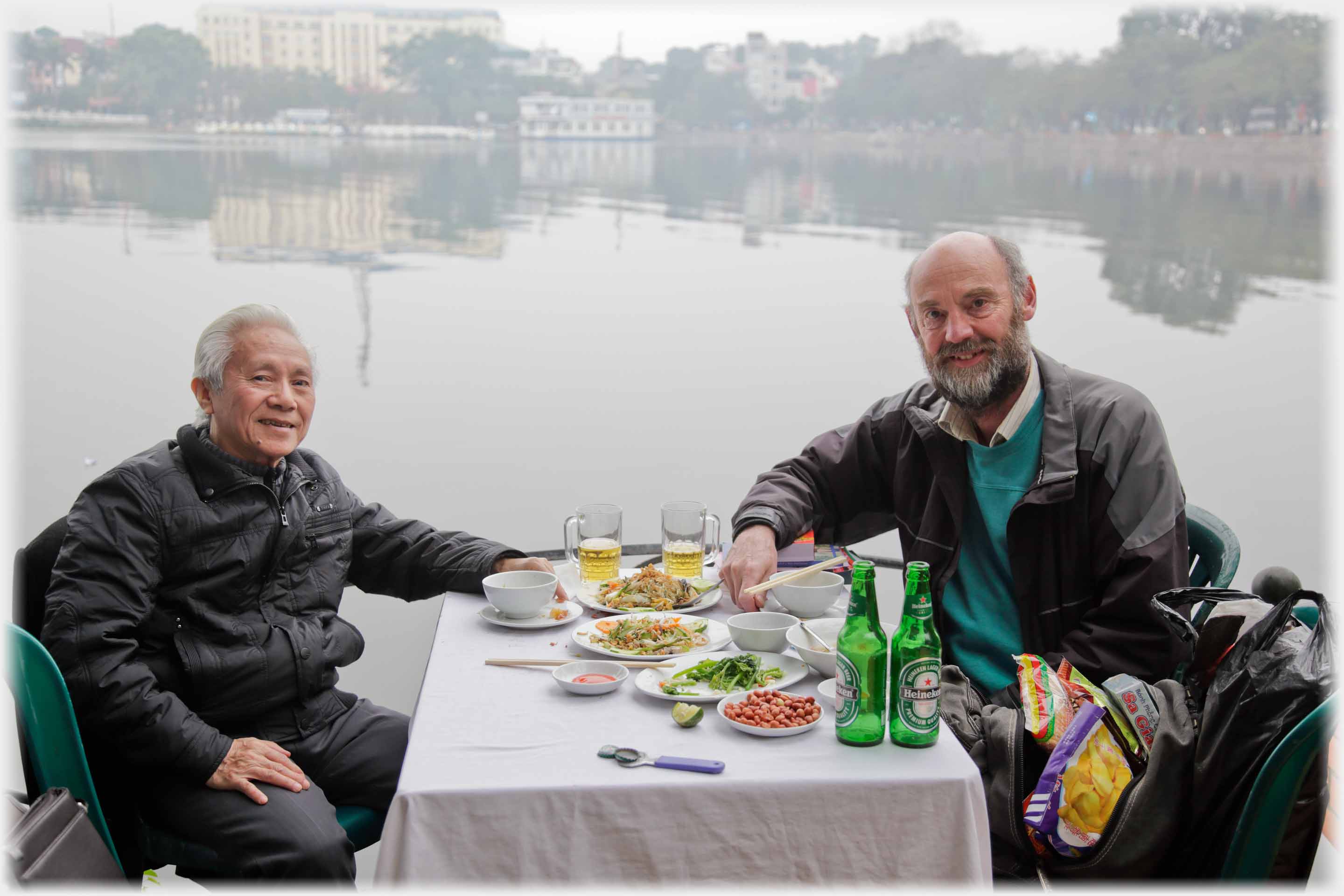 Two men at table with remains of meal by lake.