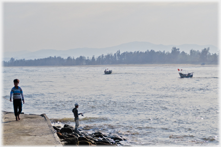 Man with rod at estuary's side, two small boats going downstream.