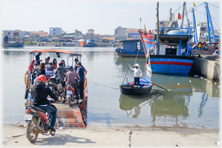 Motorbikes approaching and on the ferry, rower standing on her boat nearby.