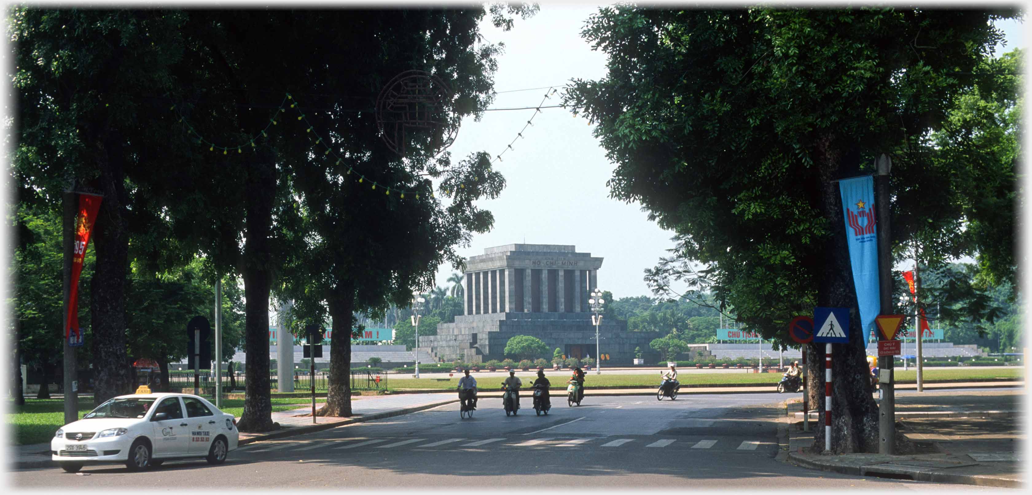 Mausoleum dominating open square seen from road between trees.