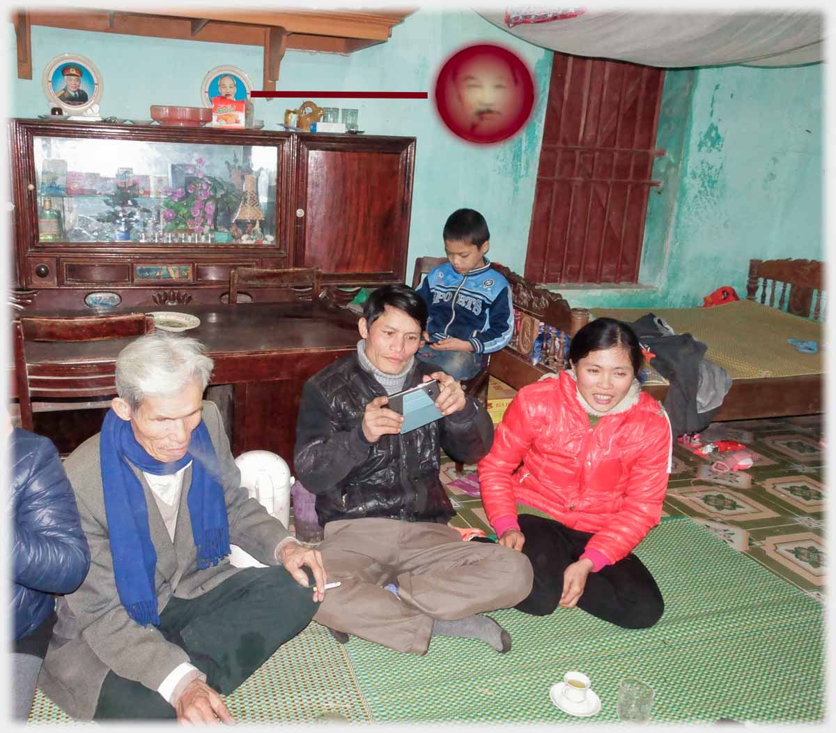 Family sitting on mats in house reception room, altar behind, small face of Ho indicated.