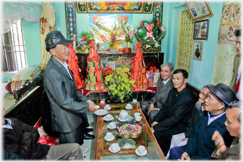 Man standing addressing seated group beside altar which is covered in wedding gifts.