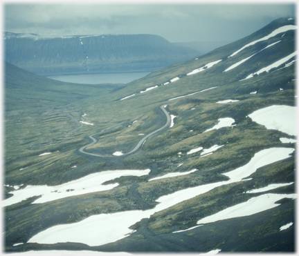 A road snaking down to a fjord far below with patches of snow around.