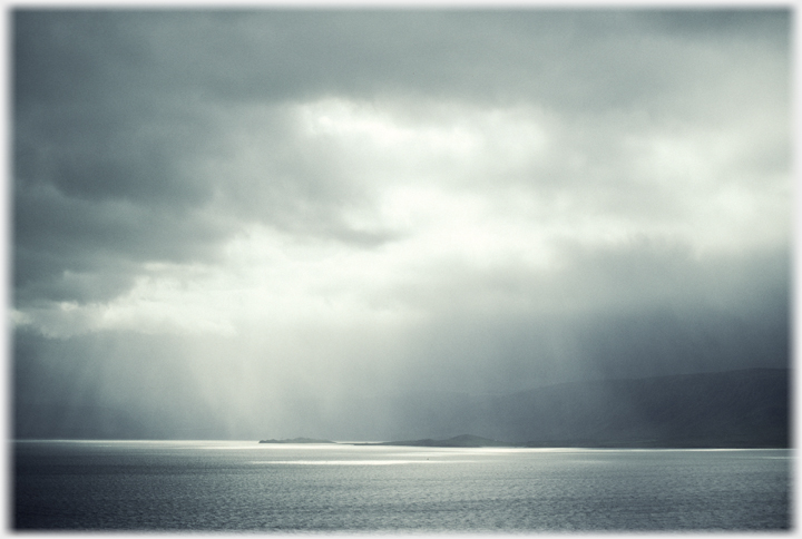 Light in shafts through cloud onto the sea near a low island.