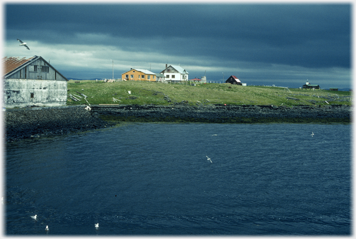 Low black clouds reflected in the sea
	 	with building for fish processing in the foreground and houses beyond.