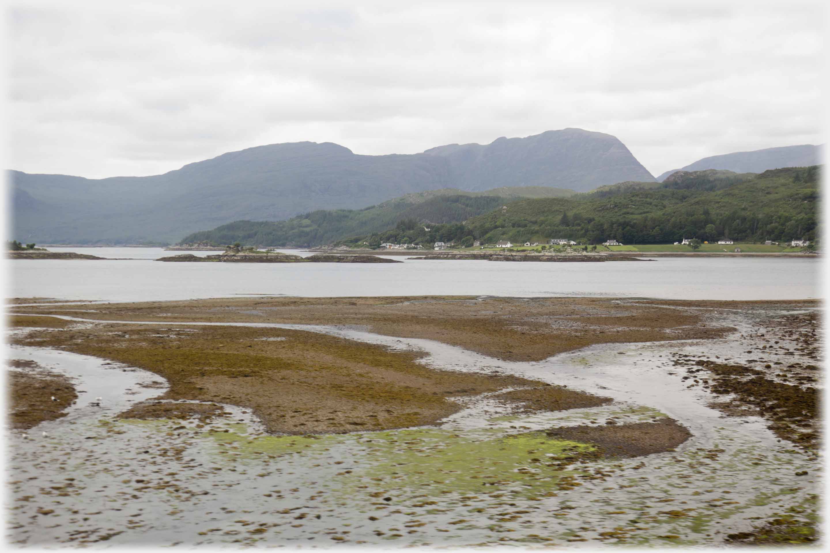 Foreground of shingle and rivulets, scattered houses on far shore, big hills rising beyond.