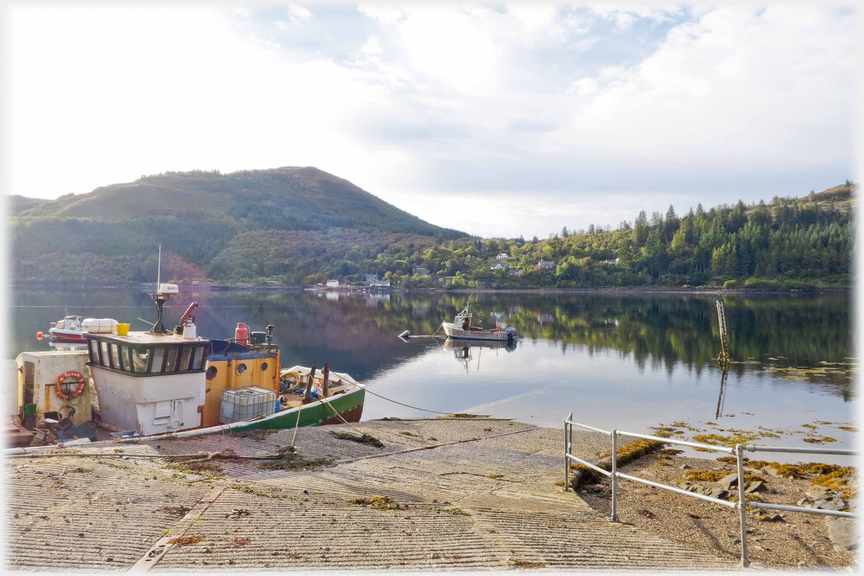 Ferry ramp into loch, fishing boat tied up to it, treed slope across the water.