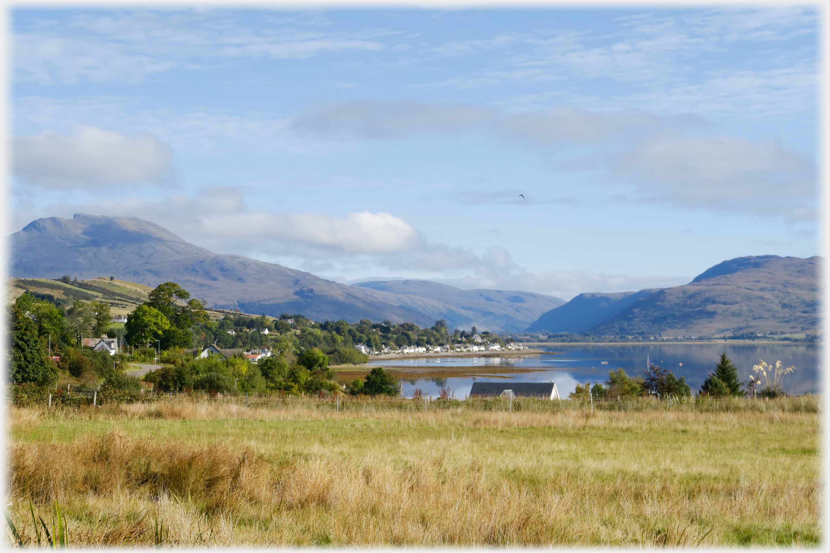 Sunlit scene of houses at lochside aand hills beyond.