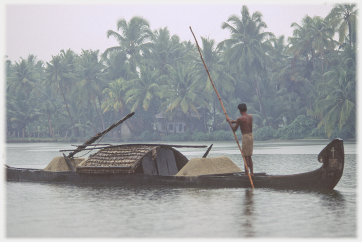 Man punting long boat with cabin and piles of sand.