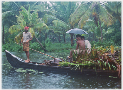 Two men sitting under umbrella on palms in boat, one standing in prow of boat.