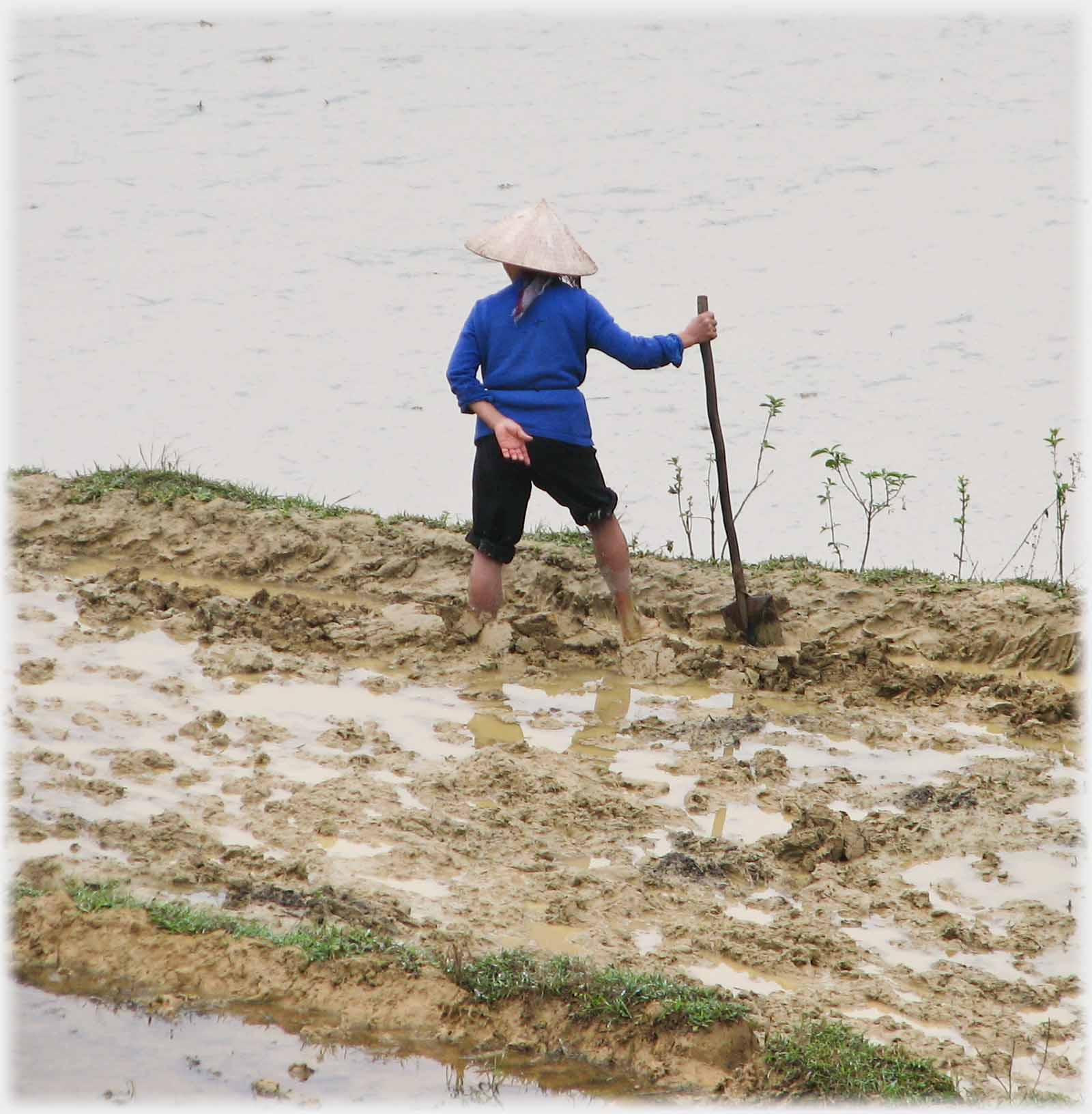 Back of woman standing in mud holding shovel.
