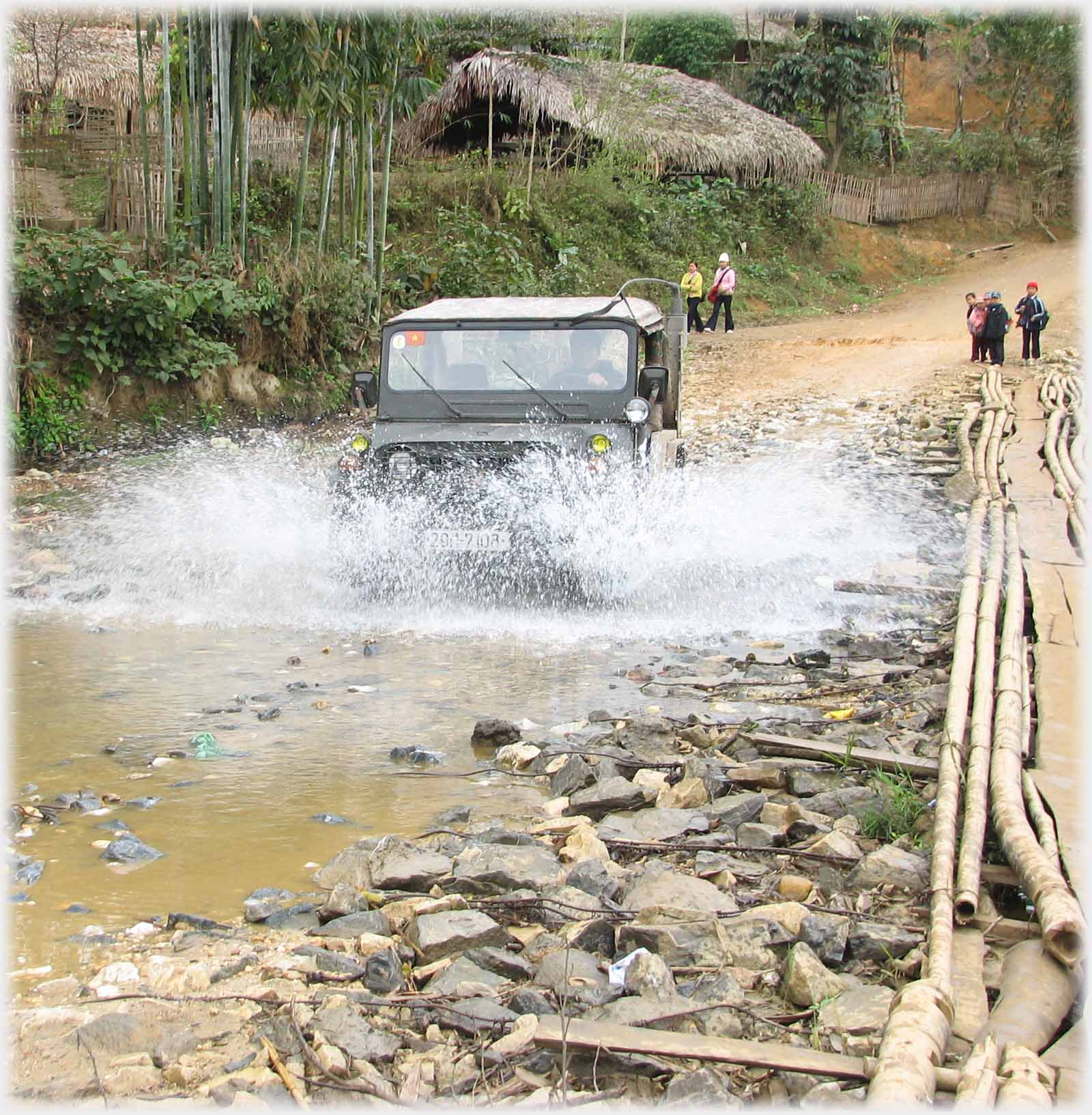 Jeep raising water through shallow stream, minimal bridge at one side.