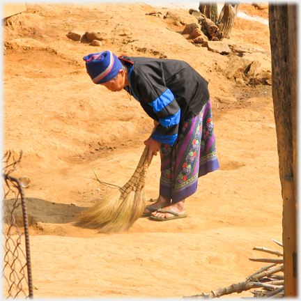Woman in local garb sweeping dusty earth.