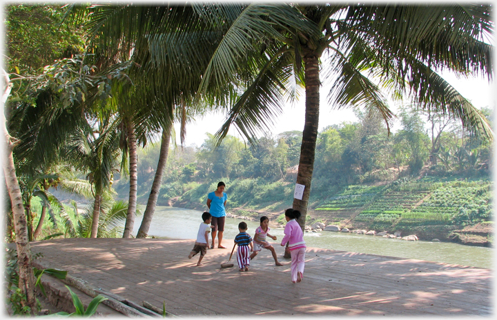 Children playing, woman watching, river and vegetable rows beyond.