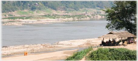 River Mekong shore with two monks and cafe on bank above.