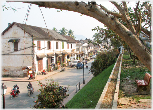 A street lined with shops, a bank to the right with seat nearby.