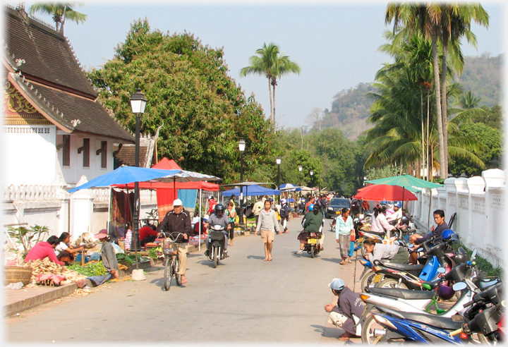 Street with vendors along left pavement, motor-bikes to right, and palm trees around.