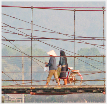 Two women walking over bridge, one with long hair pushing a cycle.