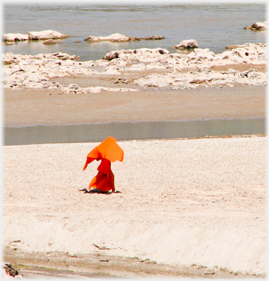 Monk striding near river shore with robe flowing over his head.