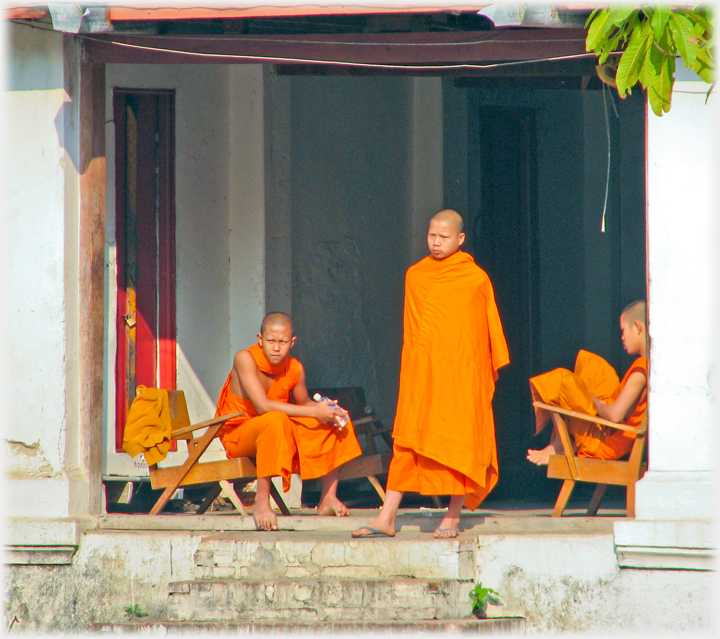 Monk stands looking out, robe swathing him, in open sided room, two others sit on either side.