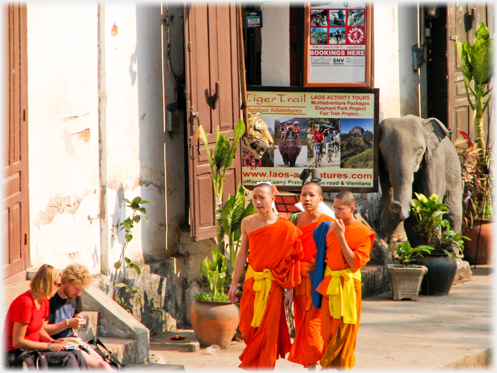 Three monks pass sitting tourists with tour agency and model elephant behind them.