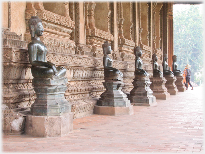 Line of Buddha statues on plinths against ornately carved wall.