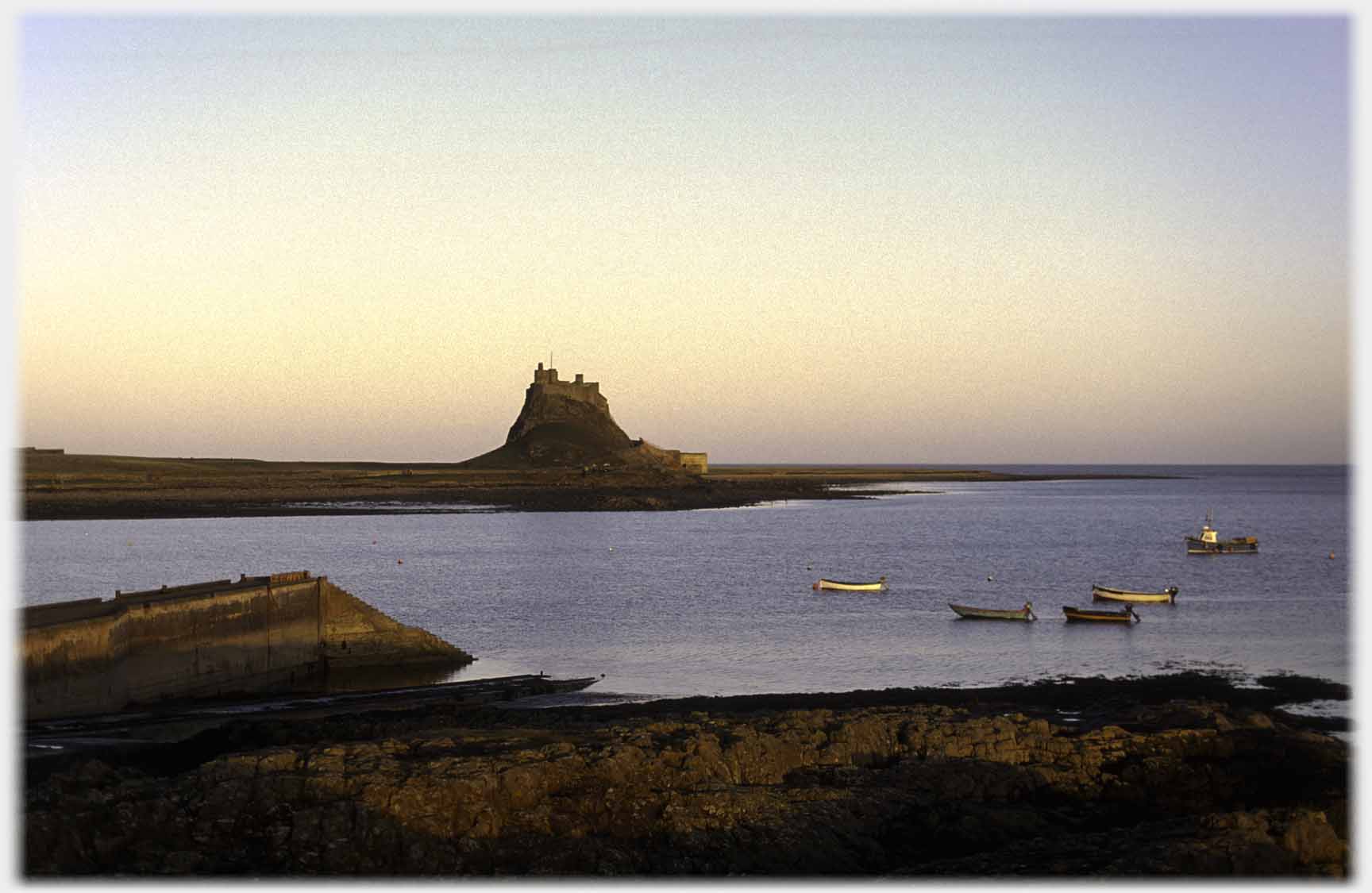 Castle on hillock, sea in foreground with five boats anchored, shore rocks,light nearly gone.