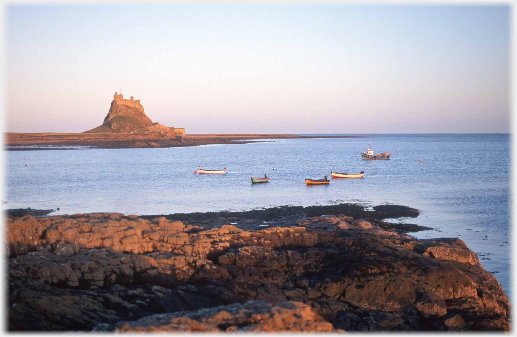 Castle on hillock, sea in foreground with five boats anchored, shore rocks.