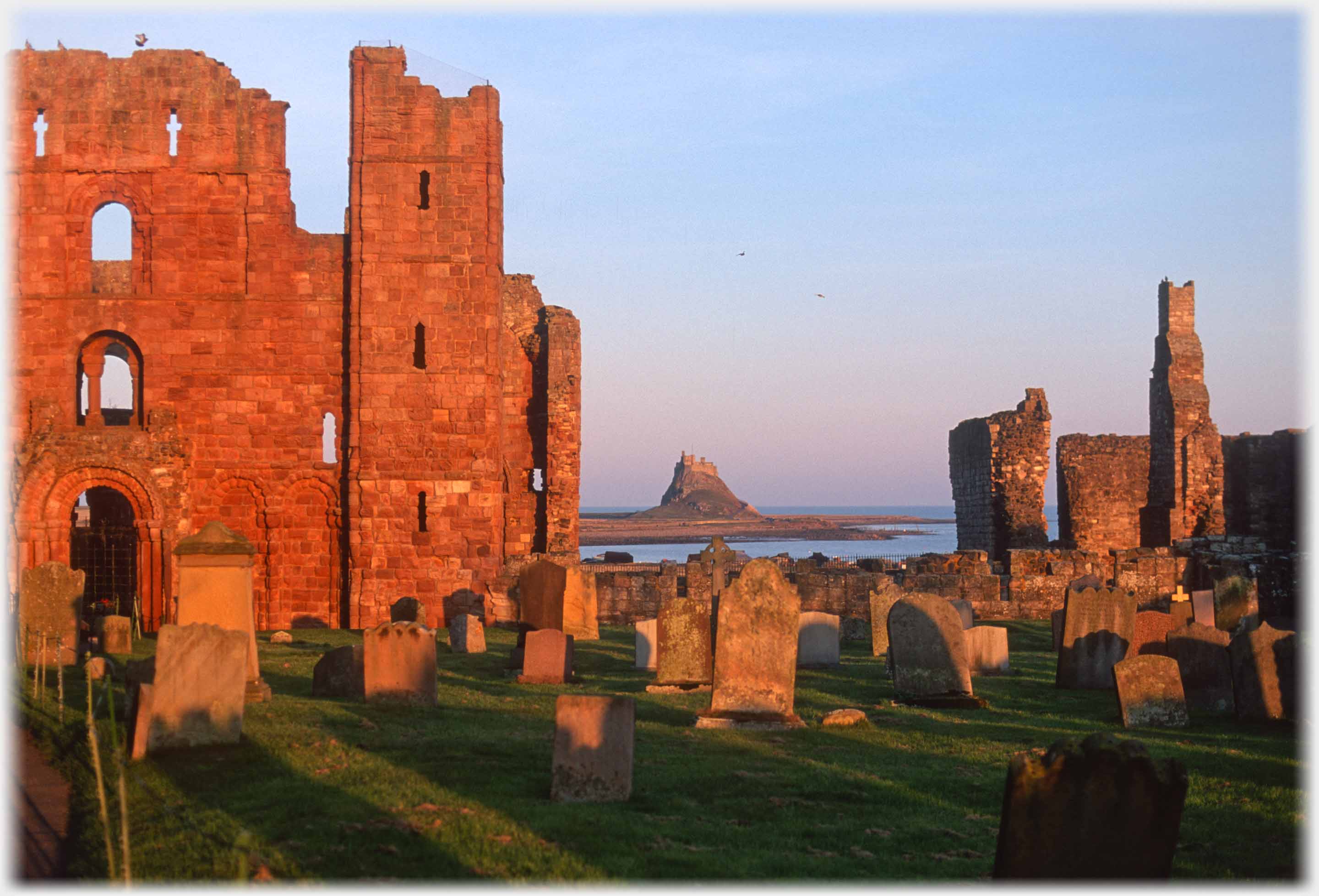 View over gravestones, by ruin, of castle across water.