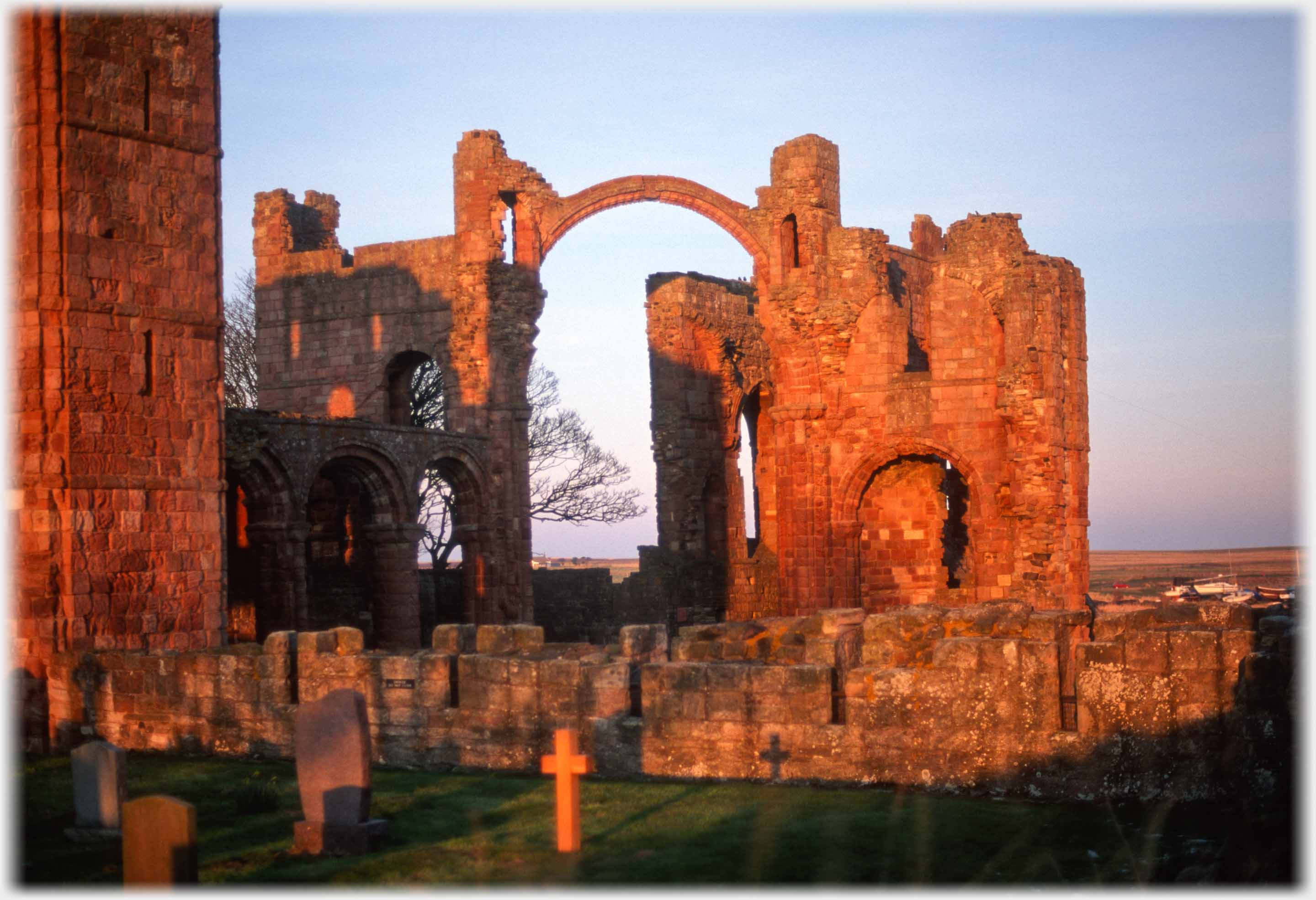 Walls of ruined church supporting high slender arch, gravestone cross and its shadow in foreground.