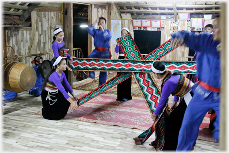 Three pairs of women holding holding broad coloured bands forming a six-pointed star.