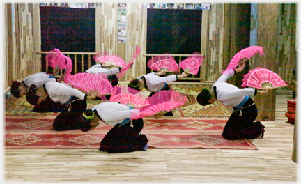 Six women with pink fans kneeling  heads towards the ground.