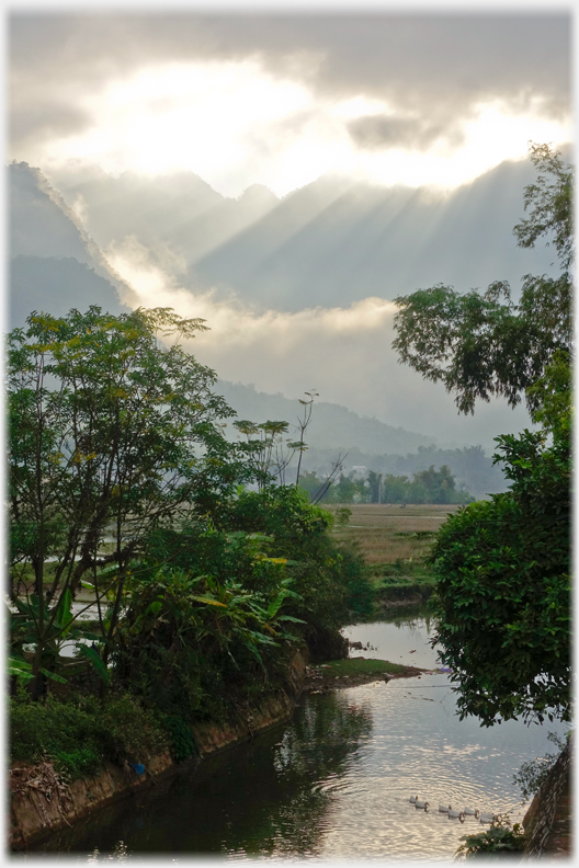 Steep hills with sun bursting over them and a river in the foreground.