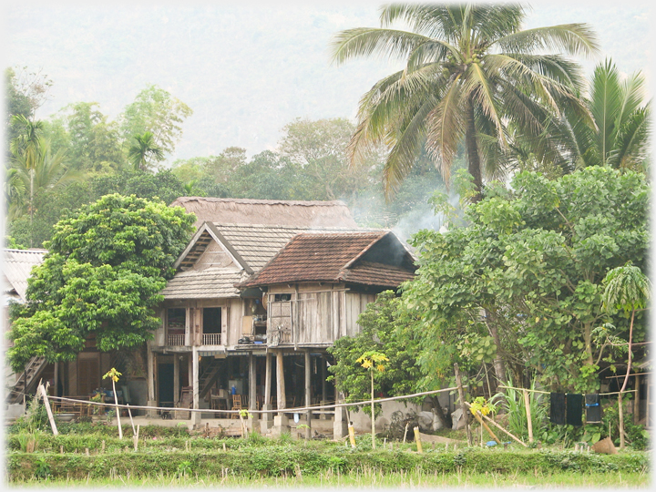 Thai stilt house among trees with smoke hanging.