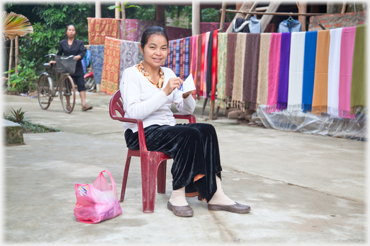 Young woman sitting sewing with line of cloths for sale behind her.