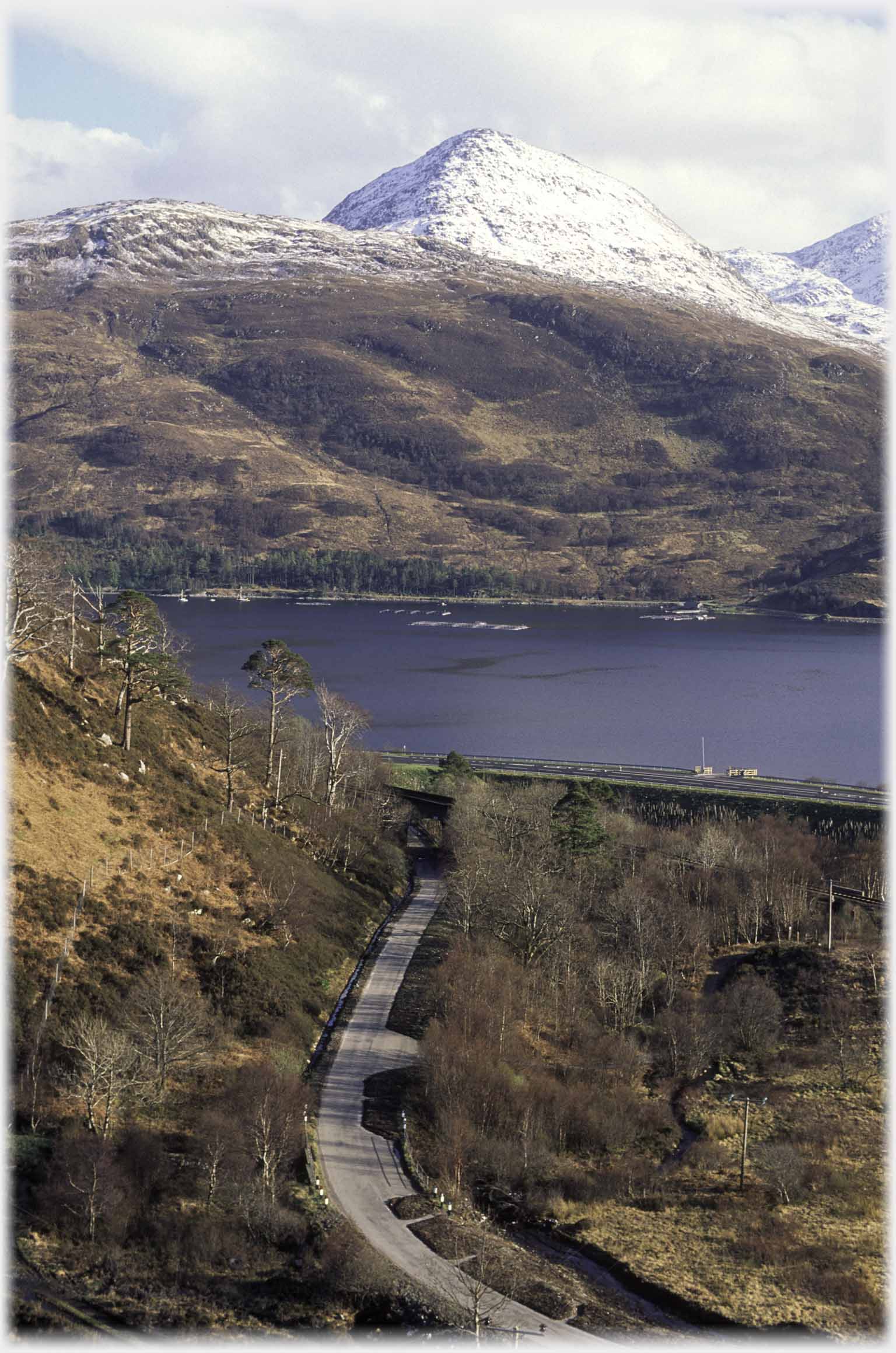 Early spring view of hill covered in snow foreground snow-free with road and rail line.